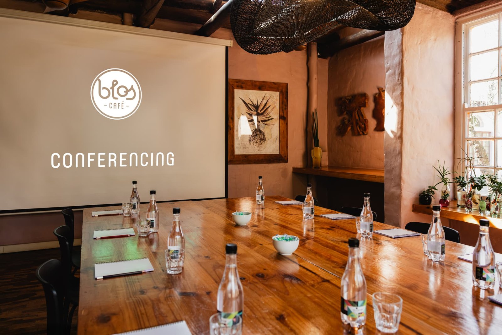 The Blos conferencing room: a long polished timber table set with notepads, pencils, water bottles and glasses, facing a projector screen showing the Blos Café logo and the word CONFERENCING