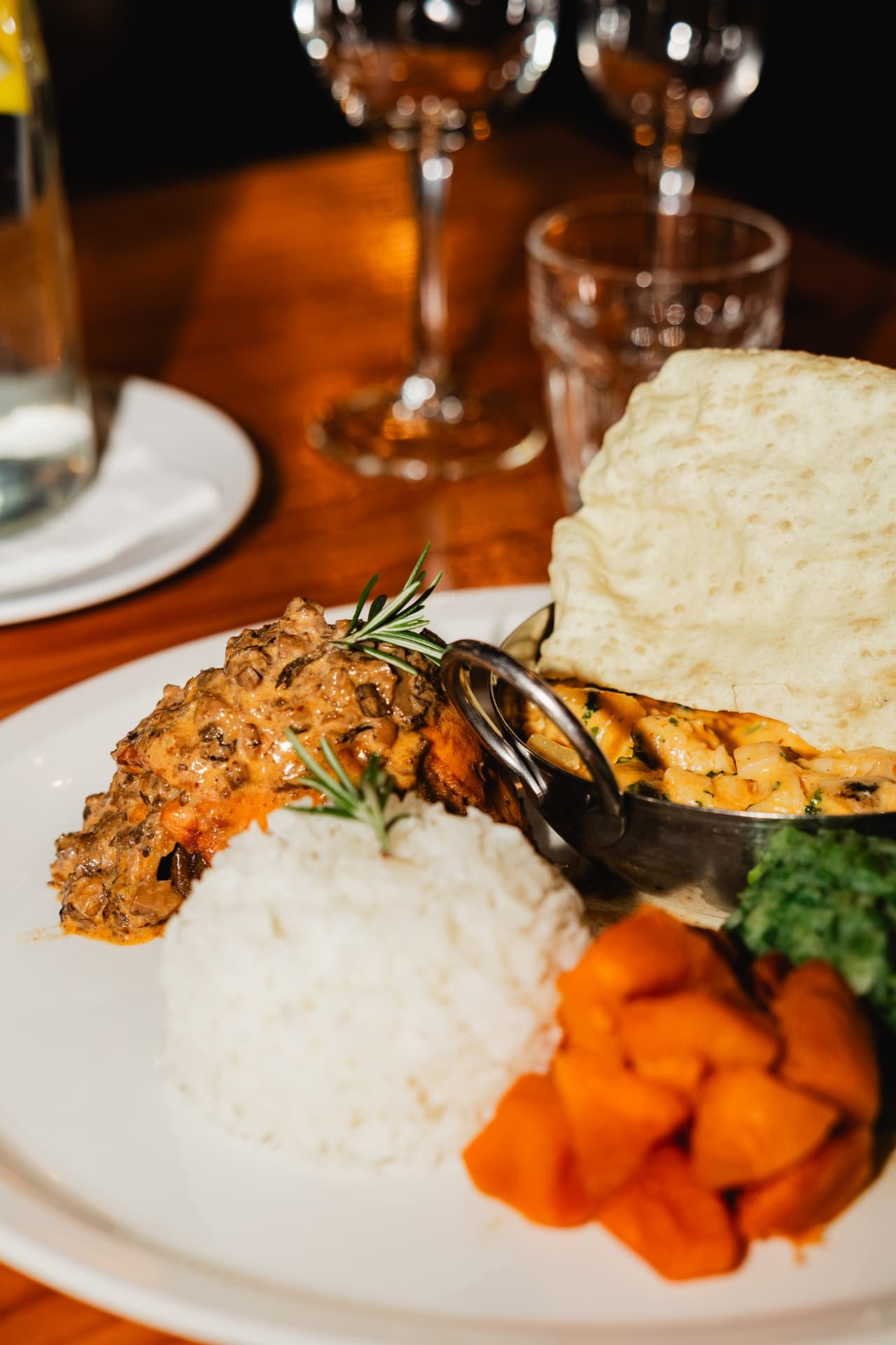 A plated Blos main course: chicken in a creamy mushroom sauce with rosemary, steamed white rice, glazed butternut, spinach, a side dish of curried vegetables and a crisp poppadom, on a candlelit table