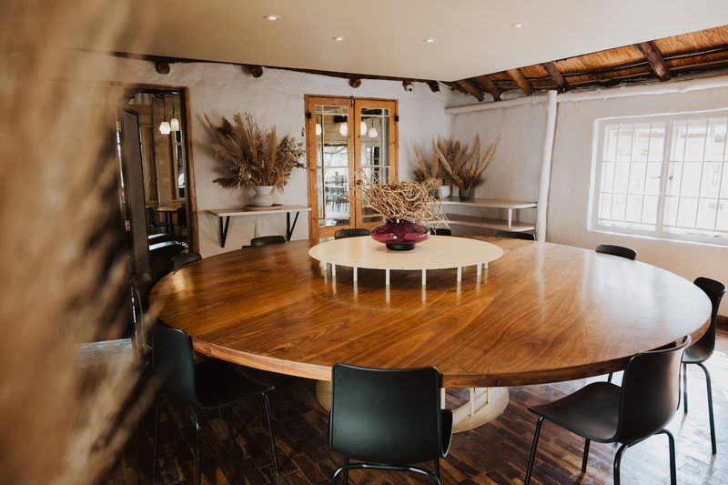The Cinema Room at Blos Café: a large round timber table ringed with black chairs beneath thatch beams, dried pampas arrangements along the walls and light from tall paned windows