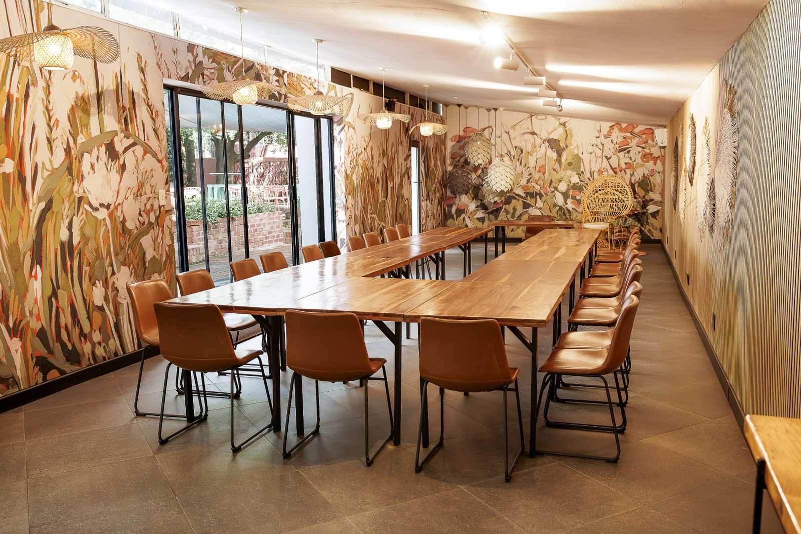 Bloot at Blos Café set with long timber tables and tan leather chairs beneath woven pendant lights, with glass doors open to the courtyard
