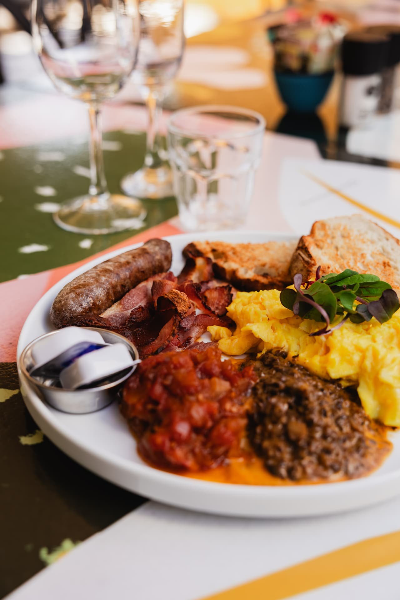 A plated Blos breakfast: scrambled eggs, three rashers of bacon, boerewors, tomato relish, creamy mushrooms and toast, on a white plate at a sunlit table